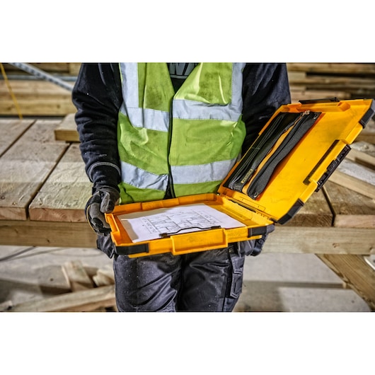 A person wearing a reflective safety vest holds an open yellow DEWALT case containing documents and tools at a construction site with wooden beams in the background.
