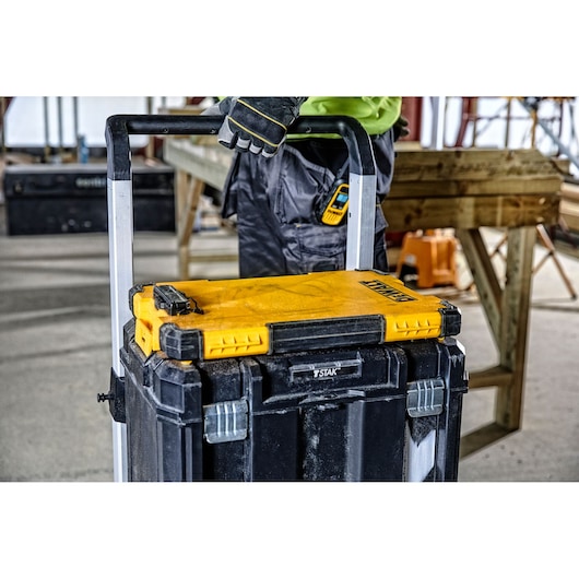 A DEWALT tool storage box with a yellow lid, mounted on a rolling trolley, is being held by a person wearing work gloves at a construction site.