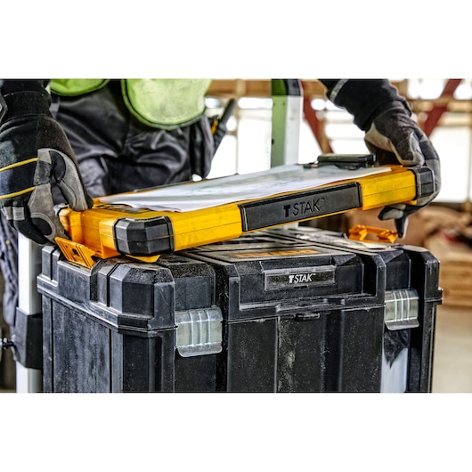 A person wearing work gloves is placing a yellow and black DEWALT T-STAK tool organizer on top of a larger black DEWALT T-STAK storage box. The scene appears to be in a workshop or construction setting.