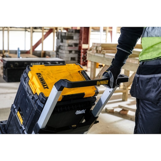A person wearing work gloves and a high-visibility shirt is pushing a DEWALT stackable tool storage trolley inside a construction site, with wooden planks and cinder blocks in the background.