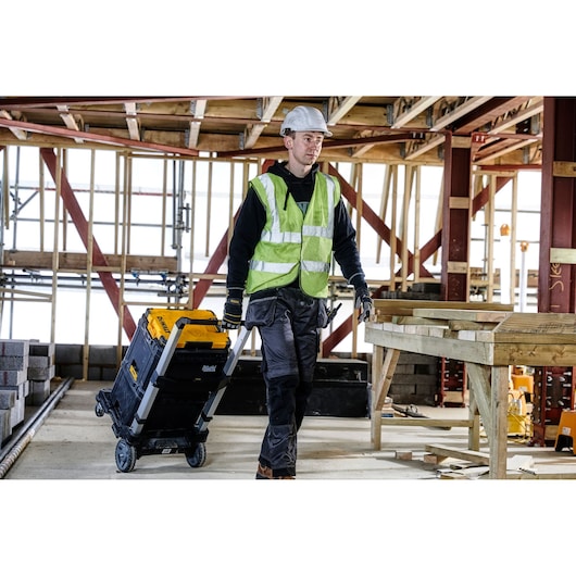 A construction worker wearing a reflective vest and hard hat is pulling a DEWALT rolling toolbox (model DWST82732-1) on a wheeled cart inside a partially constructed building site.