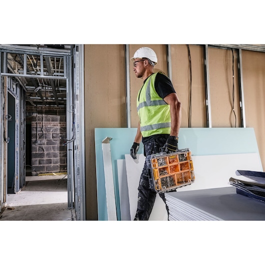 Person in safety gear carries DEWALT organizer in a construction site with drywall panels.
