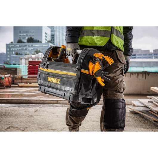Person carrying a DEWALT tool bag with tools at a construction site, wearing protective gear and gloves.