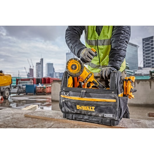 Worker grabs a DEWALT tool from a black and yellow tool bag at a construction site with city buildings in background.