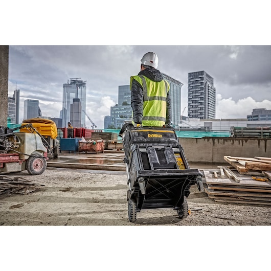 Worker on construction site pulls DEWALT DWST82990-1 tool chest, city buildings visible in background.