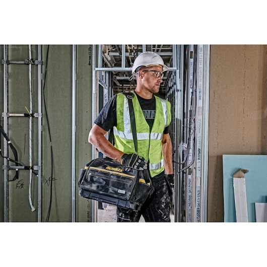 Worker in safety gear holding a DEWALT tool bag at a construction site with exposed walls and wiring.