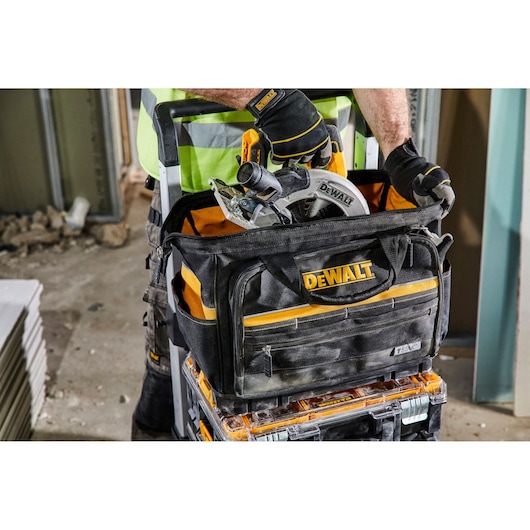 DEWALT tool bag with circular saw and tools on construction site. Person wearing gloves and safety vest handles equipment.
