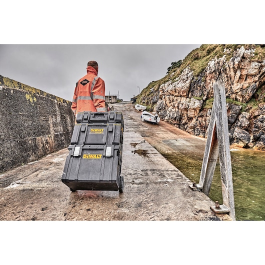 Person pulls stacked DEWALT toolboxes on wheels up a wet pier with boats and rocky background.