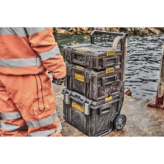 Stacked DEWALT tool boxes on wheels near water, next to person in orange workwear.