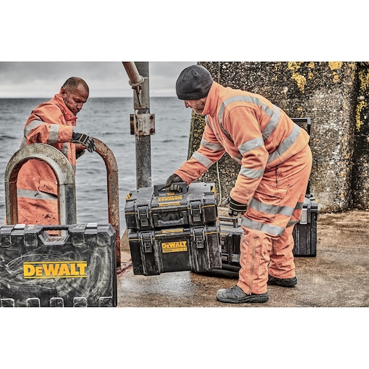 Two workers in orange suits with DEWALT toolboxes by the sea. Toolboxes are black with yellow DEWALT logos.
