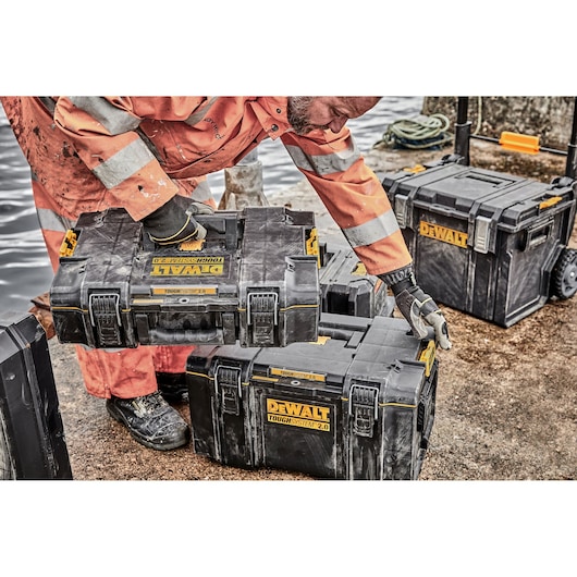 Person stacking DEWALT ToughSystem toolboxes outdoors near water, wearing orange work gear with gloves.