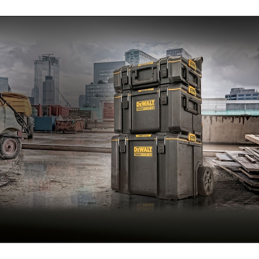 Three stacked DEWALT ToughSystem 2.0 tool storage boxes on wheels, positioned outdoors on a construction site with modern buildings in the background.