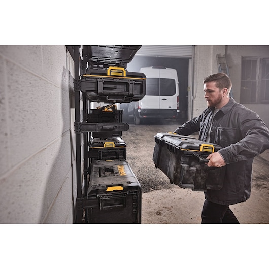 Person stores a DEWALT tool box on a wall rack in a garage with stacked black and yellow tool boxes visible.