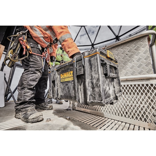 Person in work gear carries a DEWALT ToughSystem 2.0 toolbox on a construction site with metal flooring.