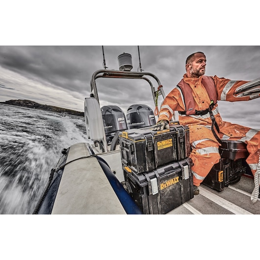 DEWALT toolboxes stacked on a boat, next to a person in orange workwear, with water and cloudy sky in the background.