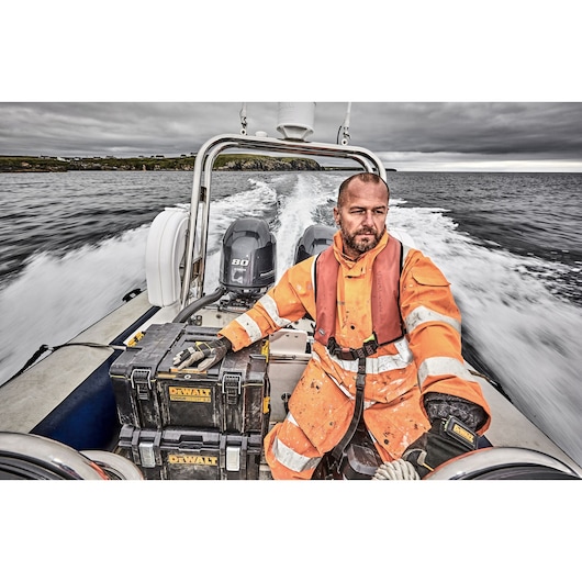 Person in orange safety gear on a boat with two DEWALT toolboxes, water and land in the background.