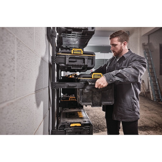 Person places a DEWALT tool box onto a storage rack in a workshop. Several black and yellow tool boxes are visible.