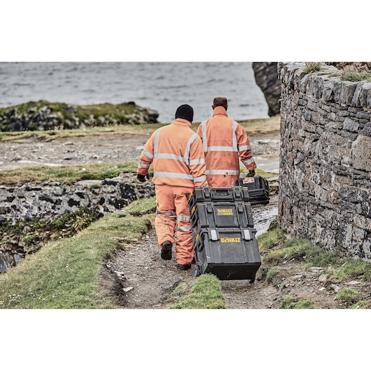 Two workers in orange suits pull DEWALT toolboxes on rough terrain near a stone wall and water.
