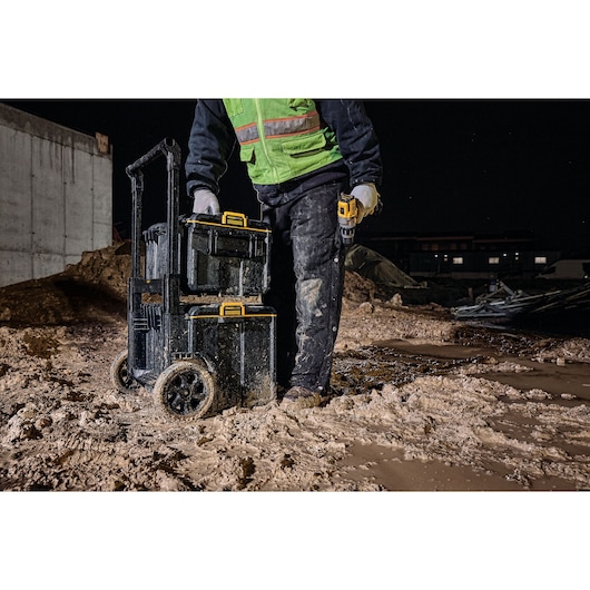 Worker with DEWALT tool boxes on wheels in muddy construction site at night.