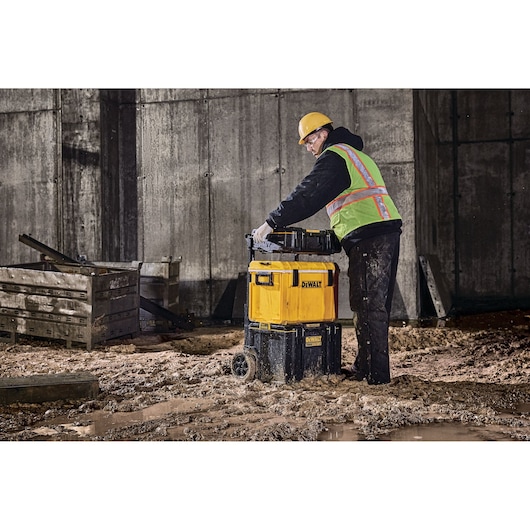 Person with safety gear uses DEWALT wheeled toolbox DWST83294-1 on a muddy construction site.