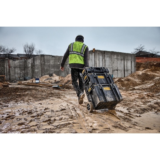 Worker pulls DEWALT tool chest on wheels through muddy construction site in rain.