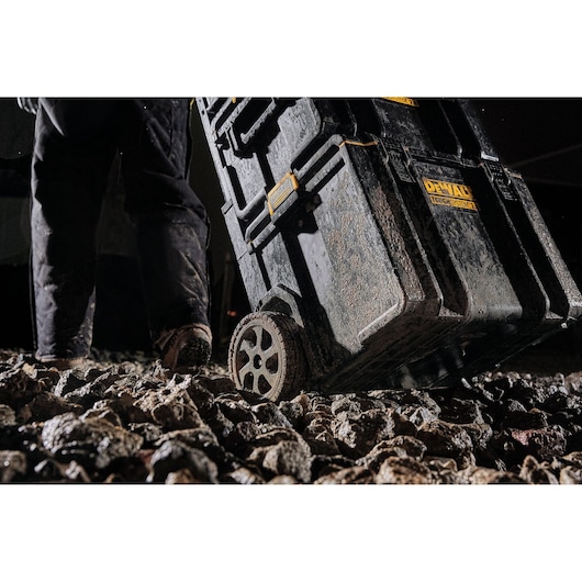 Dirty DEWALT tool chest with wheels being dragged across rocky ground by a person in work pants.