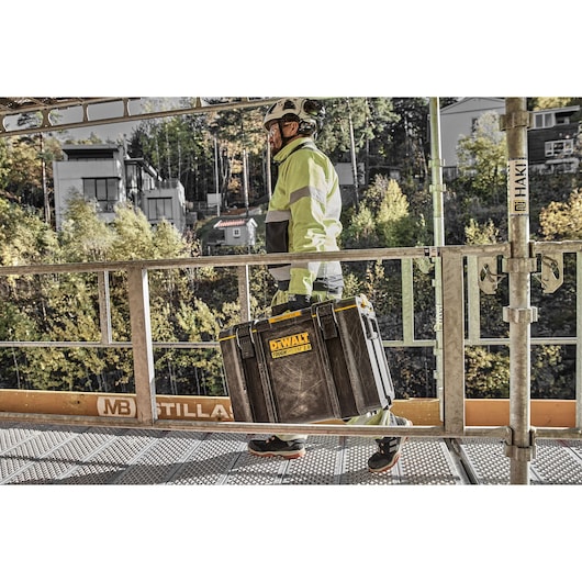 Person in safety gear carrying a DEWALT Toughsystem 2.0 toolbox on a construction site walkway.