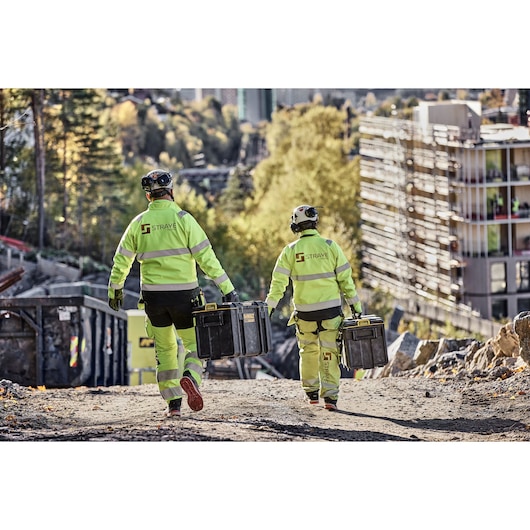 Two workers in safety gear carry DEWALT toolboxes at a construction site on a sunny day.