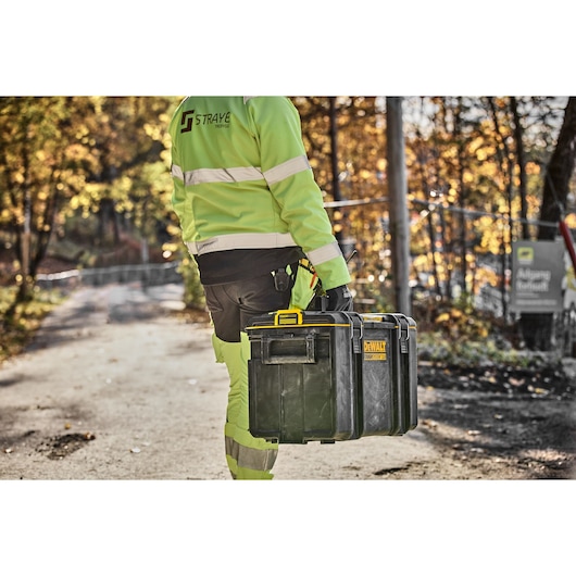 Worker in hi-vis gear carries a DEWALT DWST83342-1 black tool box on a road with autumn trees in the background.
