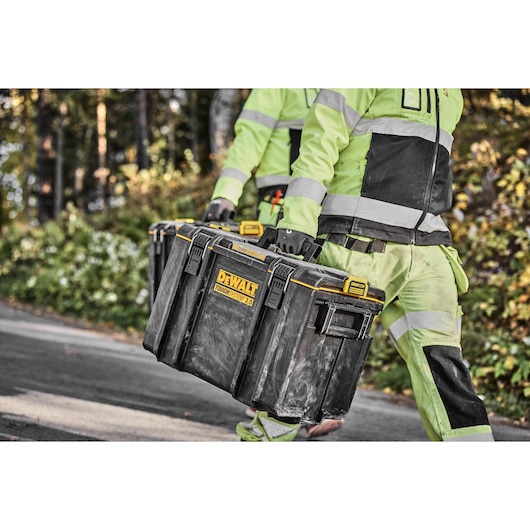 Two workers carry a DEWALT ToughSystem 2.0 toolbox outdoors while wearing high-visibility clothing.