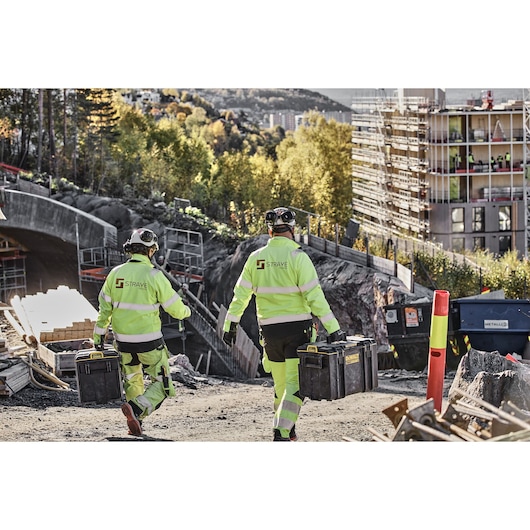 Two workers in hi-vis gear carry DEWALT toolboxes at a construction site with buildings and trees in the background.