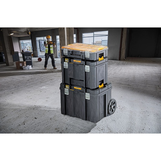 Stacked DEWALT tool boxes with wheels in a construction area, worker in background wearing safety gear.
