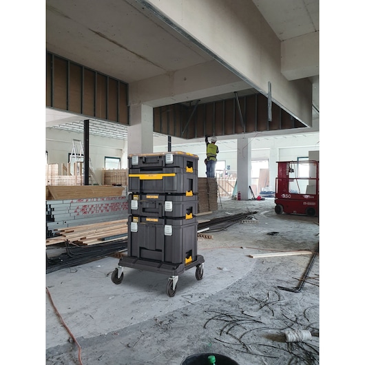 DEWALT stackable toolboxes on wheels in a construction site with materials and a worker in the background.