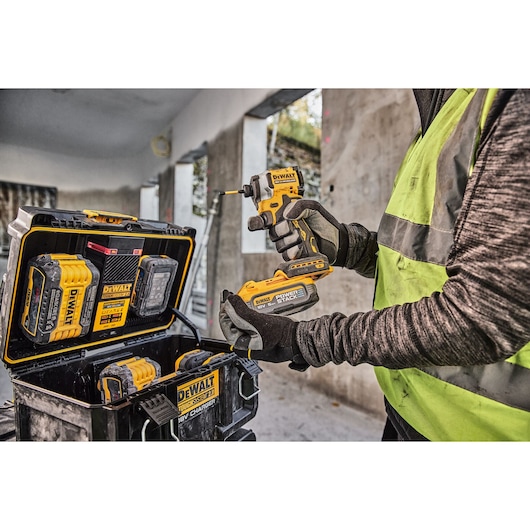 Person holding a DEWALT power tool and battery, with DEWALT tool case and batteries visible at a construction site.