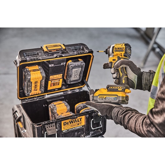 Person holding DEWALT drill, charging case with batteries and tools visible. Industrial setting.