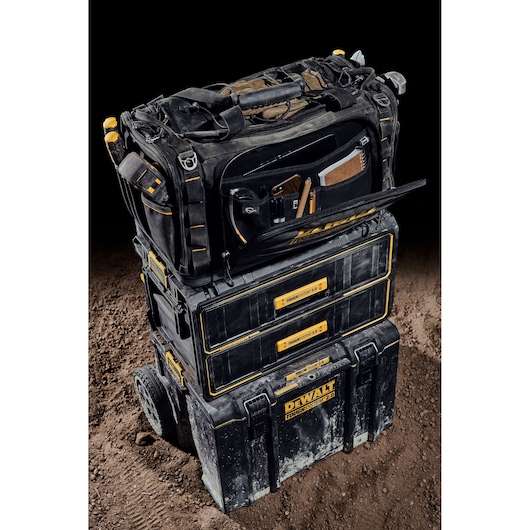 A DEWALT ToughSystem 2.0 tool storage setup is shown, consisting of three stacked black and yellow modular toolboxes. The top bag is open, revealing organized compartments with various tools and accessories. The setup is placed on a dirt surface with a black background.