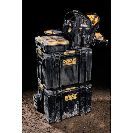 Three stacked DEWALT ToughSystem 2.0 toolboxes are shown, with a black tool backpack and a transparent organizer box on top. The toolboxes and backpack display visible dirt, suggesting use on a construction site.