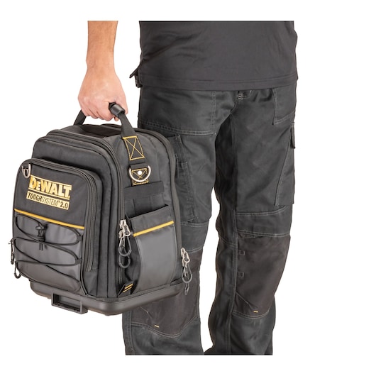 A person wearing black work trousers is holding a DEWALT Toughsystem 2.0 black tool bag with multiple zippered pockets and yellow detailing.