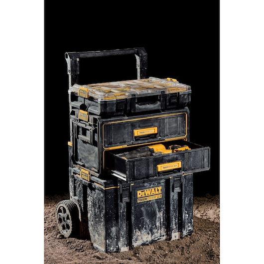 A DEWALT ToughSystem 2.0 rolling tool storage unit is shown on a dirt surface. The stackable black and yellow unit includes three compartments, with a top organizer, a middle drawer containing tools, and a large base bin. The handle is extended for transport, and the storage unit shows signs of dirt and jobsite use.