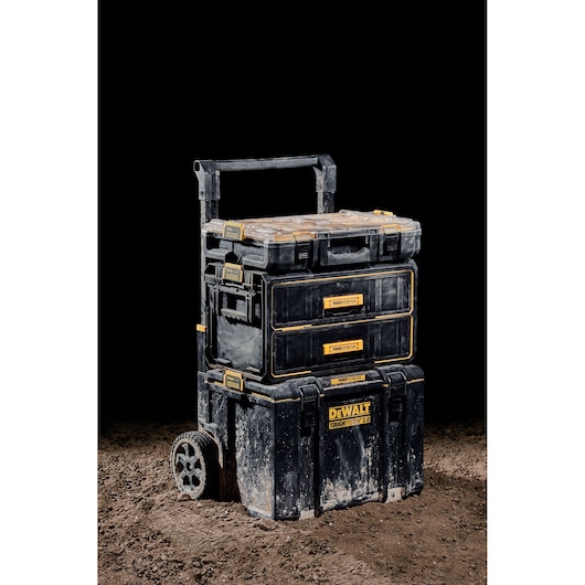 A rugged DEWALT rolling tool storage system with three stacked black and yellow toolboxes, shown on a dirt surface with a black background. The toolboxes have the DEWALT logo and are designed for professional use.