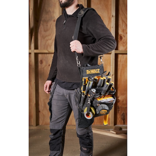Person carrying a DEWALT tool bag with various hand tools inside, standing in front of a wooden wall.
