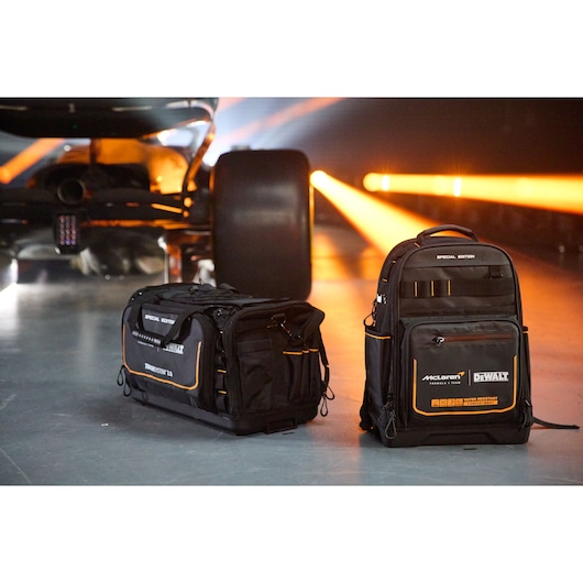 Two DEWALT McLaren tool bags, one duffel and one backpack, sit on the floor near a race car with bright lights behind.