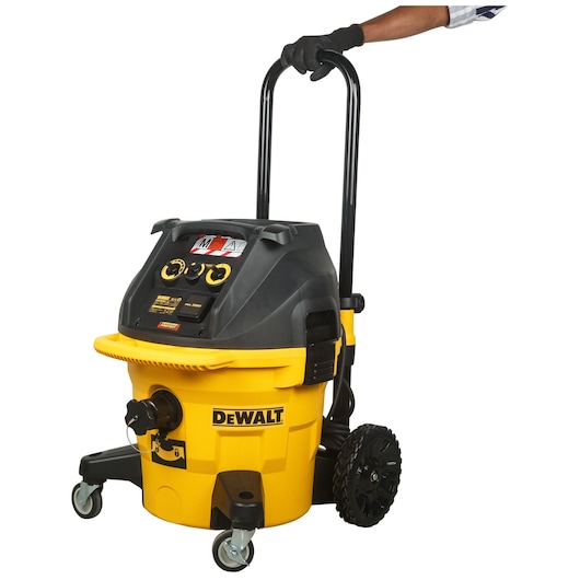 A DEWALT industrial vacuum cleaner, model DWV902M-QS, in yellow and black, equipped with wheels and a handle. It is being pushed by a person wearing a black glove.