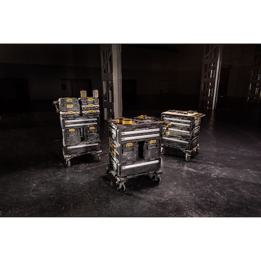 Three mobile stacks of DEWALT toolboxes with wheels are arranged in a dark industrial space. Each stack consists of multiple black and yellow storage units for organizing and transporting tools.