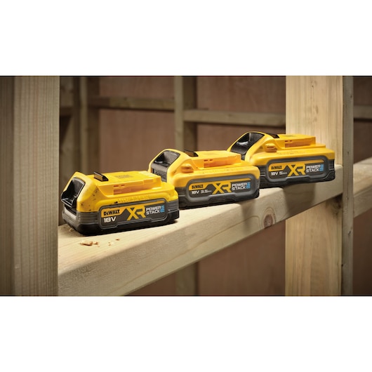 Three DEWALT XR POWERSTACK 18V batteries are placed on a wooden beam, showing their design and size differences. The background features unfinished wooden framing, indicating a construction or workshop setting.