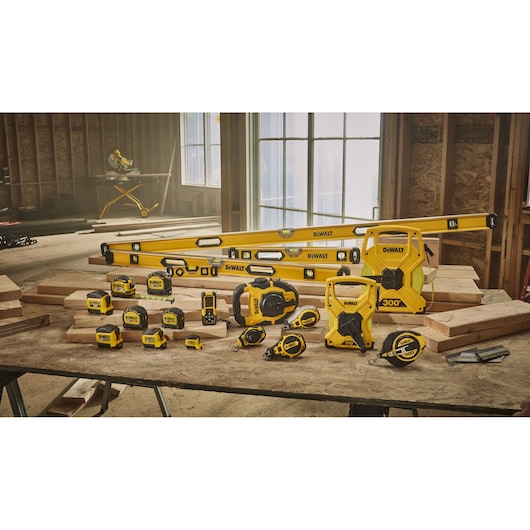 A variety of DEWALT yellow and black measuring tools, levels, tape measures, and construction accessories are displayed on a workbench in a partially built wooden structure. The tools include distance measuring wheels, tape measures, laser distance meters, and box levels.