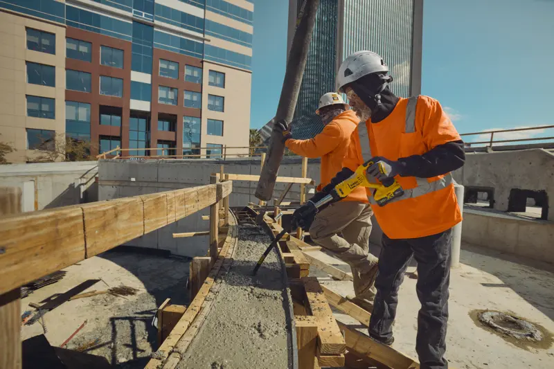 Deux ouvriers du bâtiment portant des casques de sécurité et des vestes orange haute visibilité travaillent sur un chantier. L'un utilise un outil de vibrateur à béton DEWALT sur du béton fraîchement coulé, tandis que l'autre dirige un tuyau à béton. Des bâtiments de bureaux modernes sont visibles en arrière-plan.