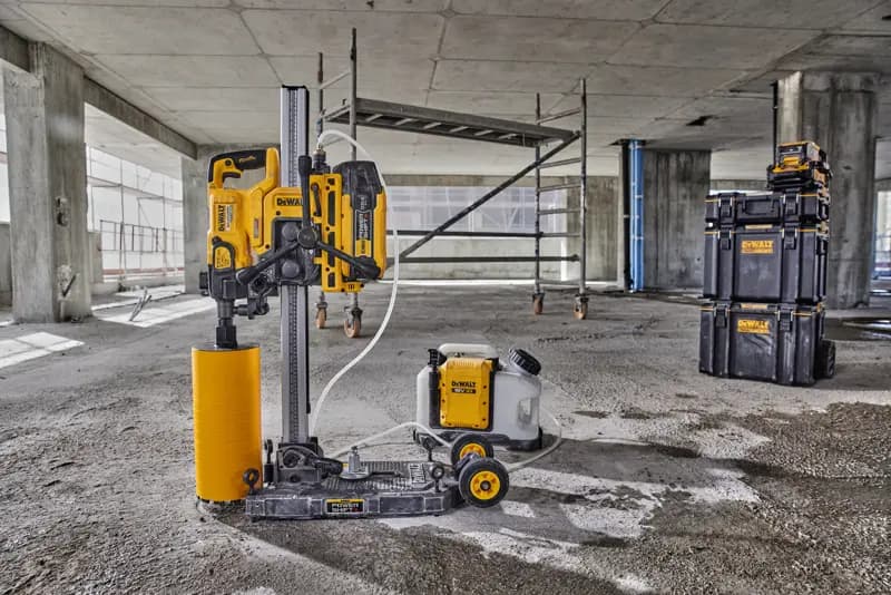 Une machine de forage de carottes DEWALT avec un accessoire de foret cylindrique jaune et un réservoir d'eau connecté est placée sur le sol d'un chantier en béton. À l'arrière-plan, on aperçoit un échafaudage et des boîtes de rangement empilables de marque DEWALT.