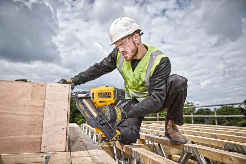 Une personne utilise un cloueur DEWALT DCN930N sur des planches en bois, sur un chantier extérieur, avec équipement de