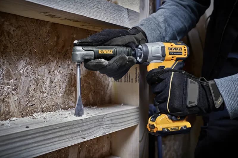 Une personne portant des gants utilise une perceuse sans fil DEWALT 12V avec un accessoire d'angle droit pour percer une planche en bois, avec des copeaux de bois visibles autour de la zone de travail.